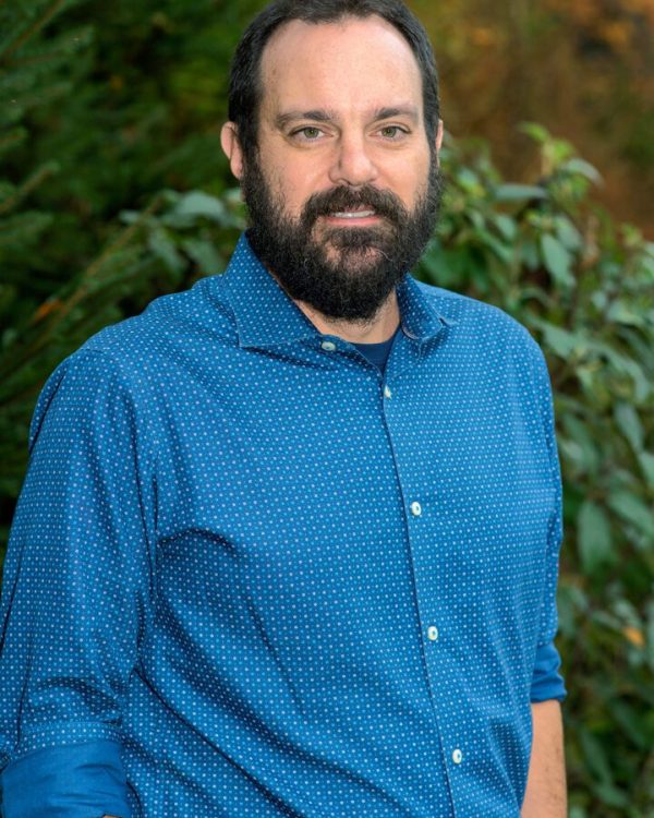 A man with beard and blue shirt standing in front of bushes.